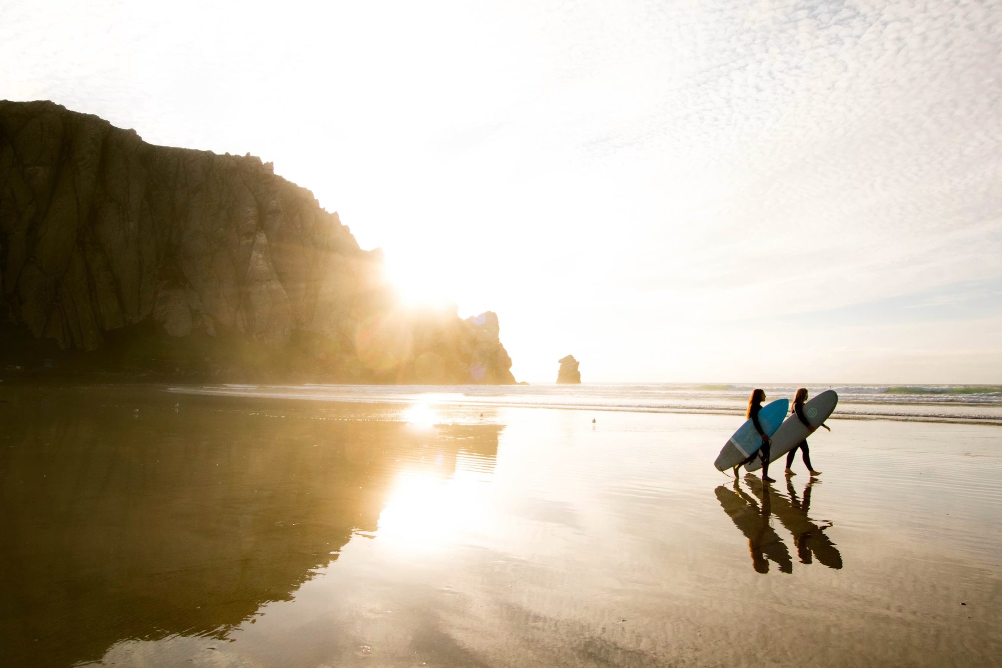 Two people walking together along the shoreline at golden hour, carrying surfboards toward the water.