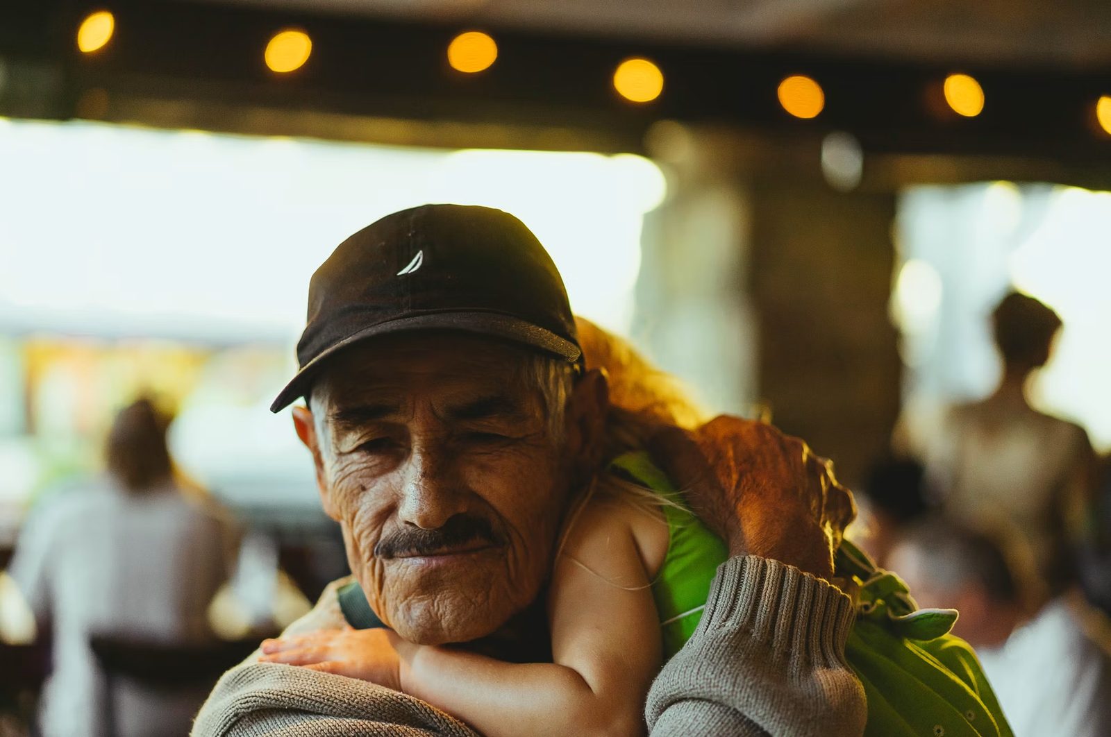 An older man in a cap holding a small child resting on his shoulder, both at peace under warm string lights.